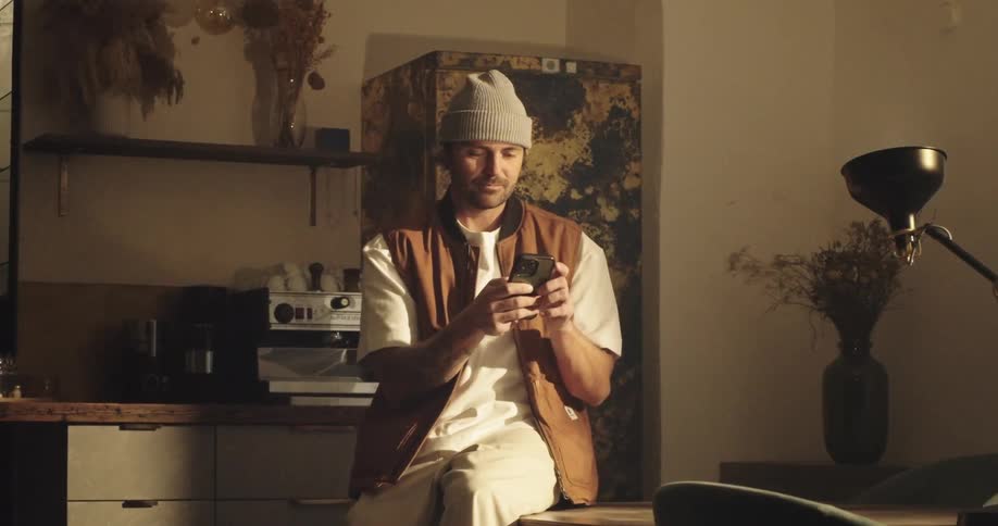 A man wearing a beanie and a brown vest sits on a kitchen table while focused on his smartphone the scene features warm atmospheric lighting that highlights the modern slightly rustic interior of the room