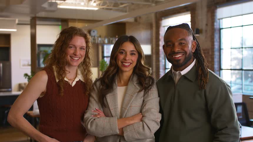 The video shows three colleagues standing together in a modern sunlit office space they transition from chatting and laughing with one another to facing the camera with warm professional smiles