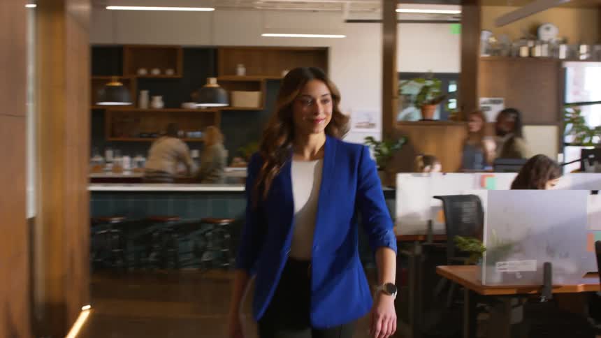 A woman wearing a blue blazer and black trousers walks through a modern office with wooden walls and a kitchen area while looking at her watch during the day