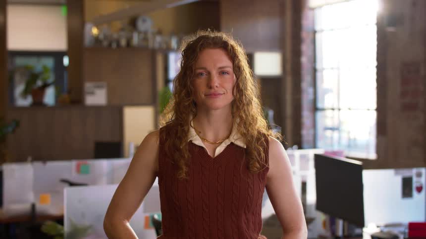 A young woman with long curly blonde hair stands inside an office area at midday smiling
