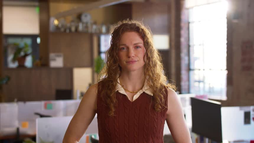 A young woman with curly red hair stands in a bright office with her hands on her hips and a gentle smile while blurred desks and computer monitors occupy the background space behind her