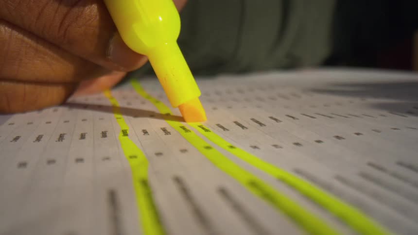A person uses a bright yellow marker to highlight lines of text on a printed document resting on a wooden surface