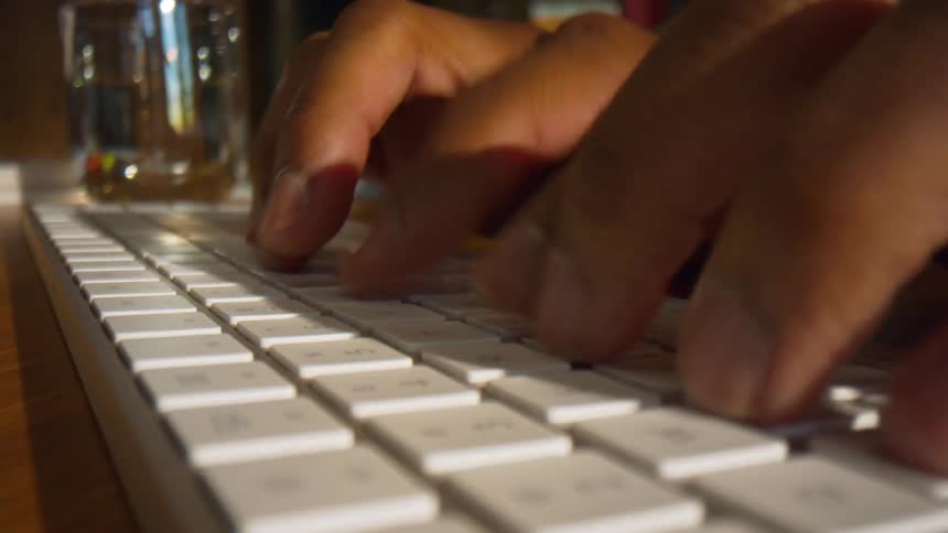 Hands type on a white keyboard on a desk with a glass in the background