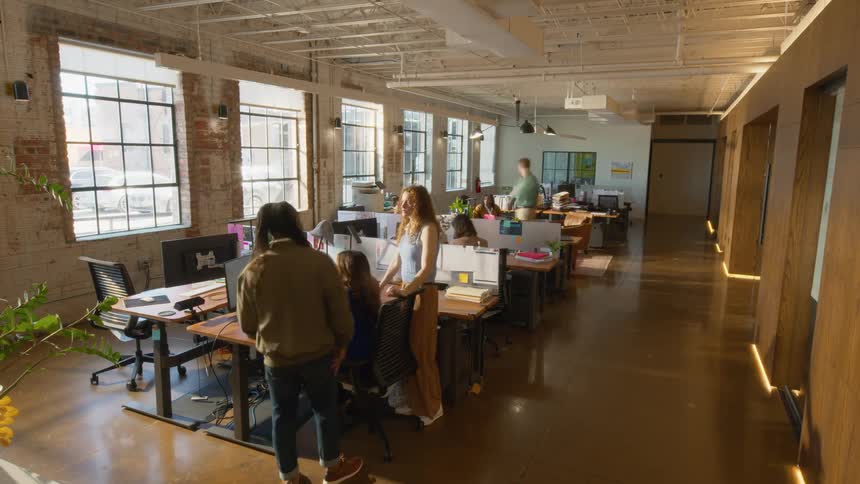 Diverse employees work and interact within a sunlit industrial office featuring exposed brick walls rows of wooden desks with laptops and large windows overlooking a city street while colleagues move through the central hallway of the workspace
