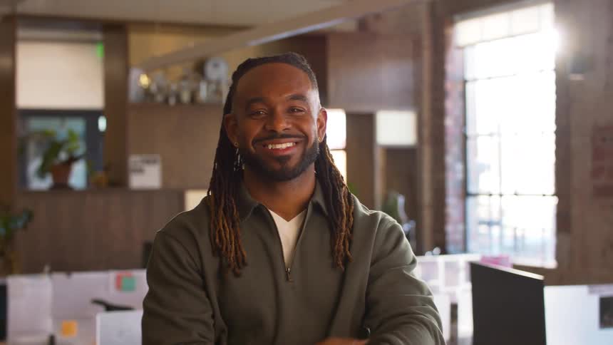 A smiling black man with long dreadlocks stands with his arms crossed in a sunlit modern office