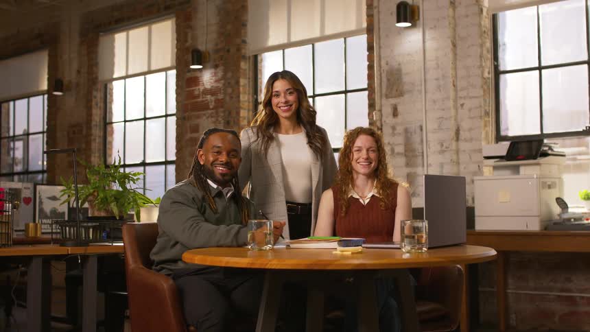 Two women and a man sit and stand around a wooden table in a modern office while smiling at the camera during the day