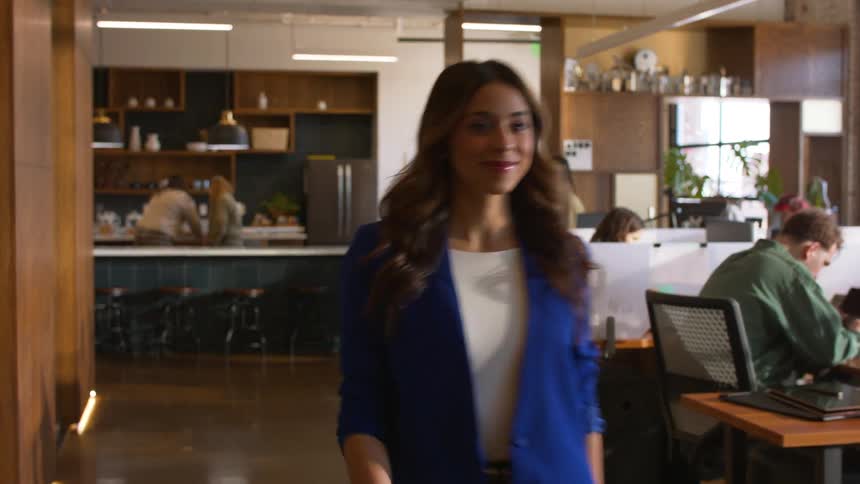 A woman wearing a blue blazer and white shirt walks through a modern open plan office while coworkers sit at desks or stand near a kitchen area in the background under warm indoor lighting