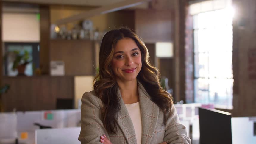The video features a smiling woman with long brown hair and a blazer standing in a sunlit office she maintains a confident pose with her arms crossed while looking directly into the camera