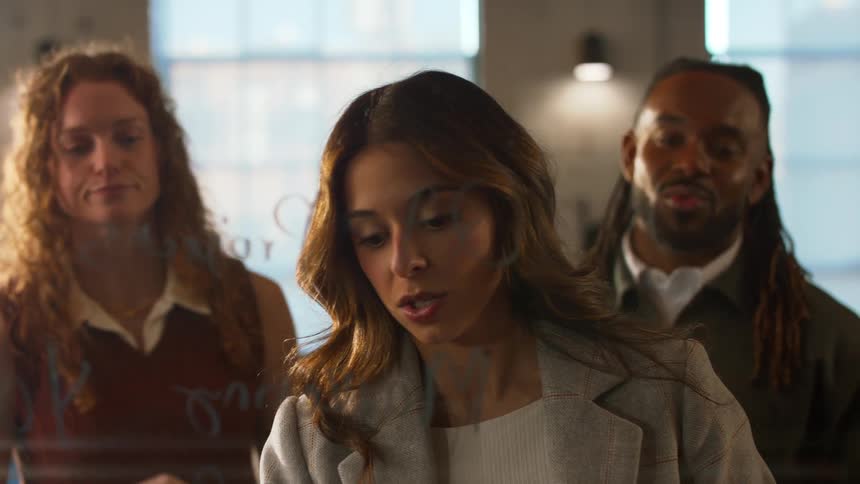 The woman in the center who is wearing a grey blazer and actively writing or pointing at notes on the glass her facial expressions shift from a smile at the start to a more serious concentrated look as she explains the content to her colleagues