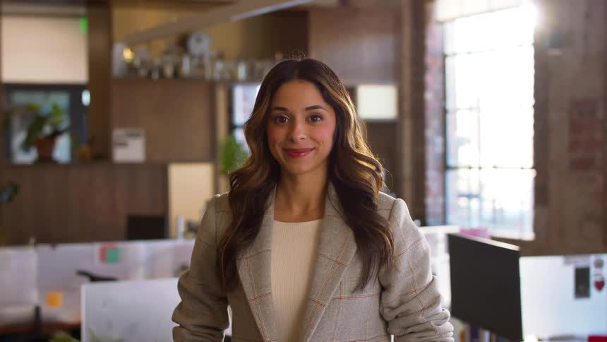 A woman with long wavy brown hair stands in a bright modern office space and smiles directly at the camera she wears a grey plaid blazer over a white top positioned in the center of the frame with a blurred workspace in the background