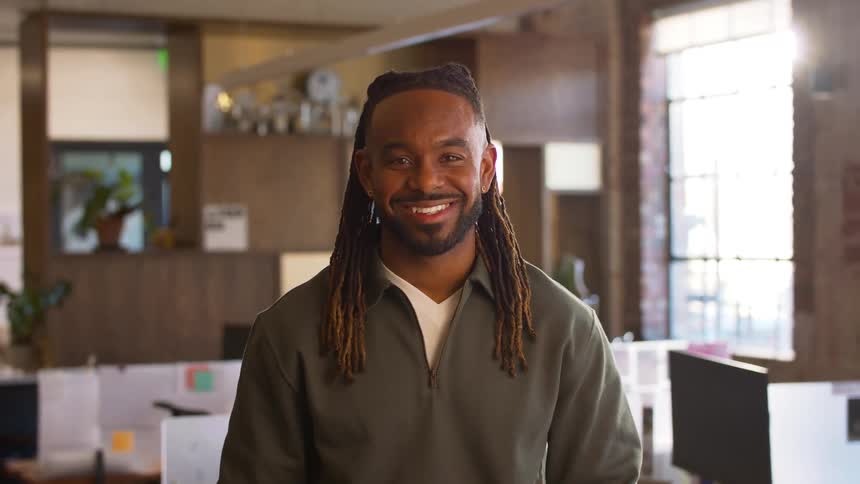 A black man with dreadlocks smiles directly at the camera in a modern office setting