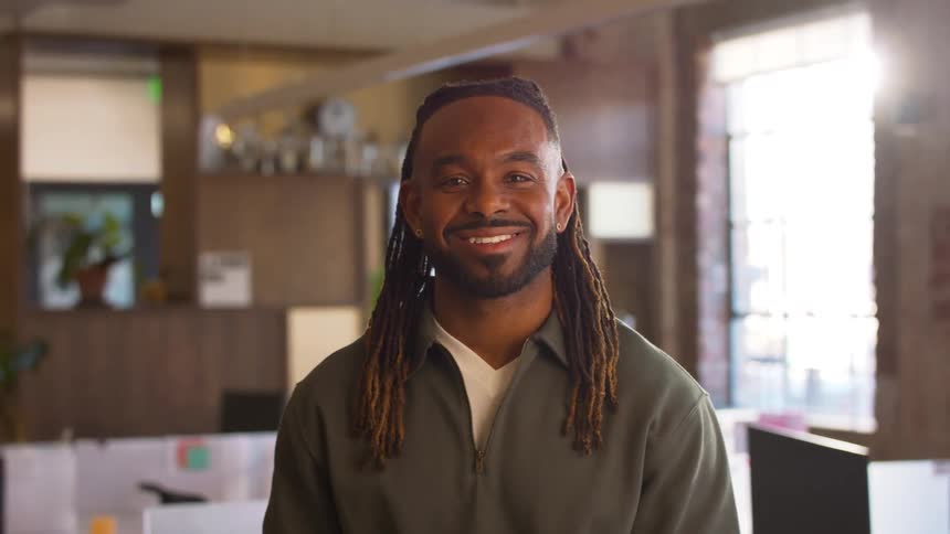 A smiling man with long dreadlocks stands in a bright modern office wearing a green pullover while blurred desks and large windows fill the background under soft natural light