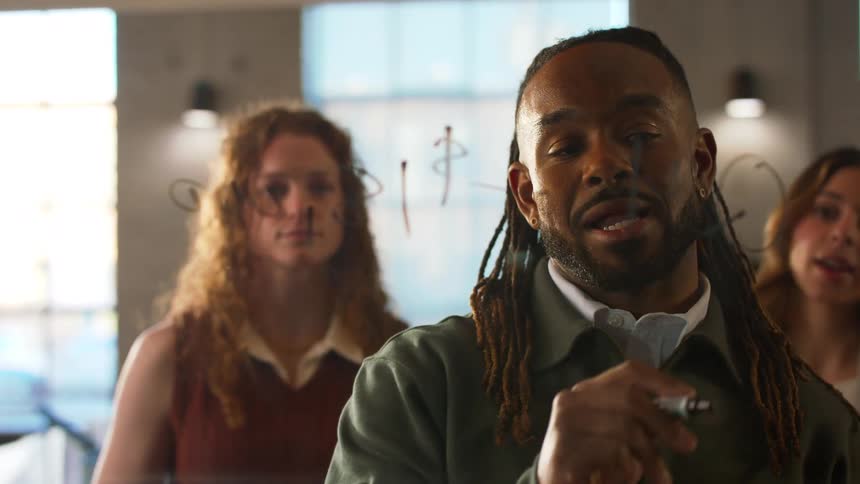 A young black man writes on a board with a marker while two young women stand behind him all communicating with one another