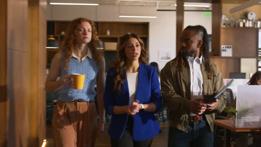 Three colleagues walk through a modern office hallway while engaged in a focused discussion. The group consists of a woman in a blue blazer speaking to her coworkers: a red-headed woman holding a yellow mug, and a black man carrying a notebook.
