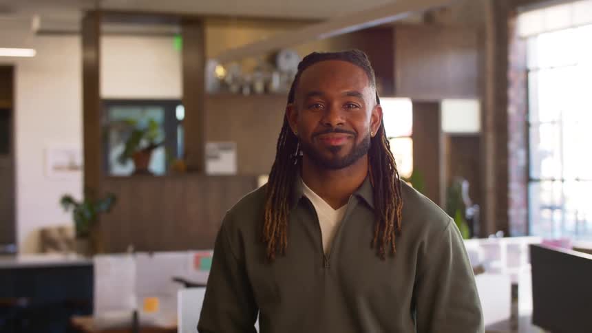 A man with long dreadlocks and a beard wearing a green pullover stands in a modern office and smiles at the camera during the day
