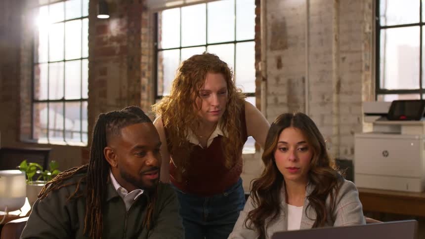Two colleagues sit at a wooden table in a bright loftstyle office focused on a laptop and scattered paperworka third woman with curly red hair approaches from behind leaning in to join their discussion as they look at the screen together