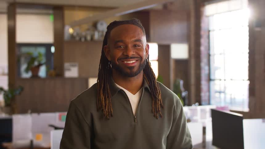 A man with long dark dreadlocks wearing a grey pullover sweater looks up and smiles at the camera while standing in a bright modern office with desks and a large window in the background