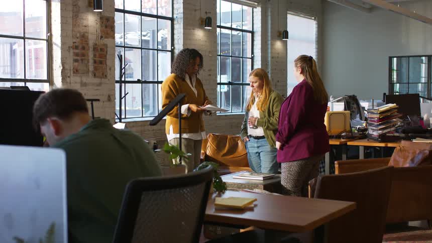 Three women talking in office with desks computers and large windows in the background