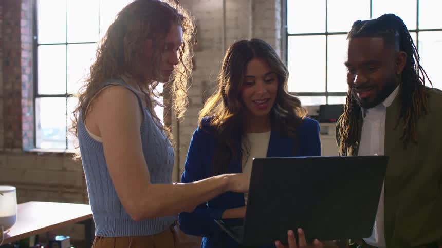 Three colleagues stand together in a sunlit industrial office while looking at a laptop screen held by a man with dreadlocks as two women gesture toward the computer and engage in a professional conversation