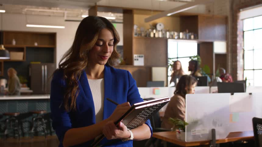 Woman in office holding tablet and pen in front of desks and coworkers working