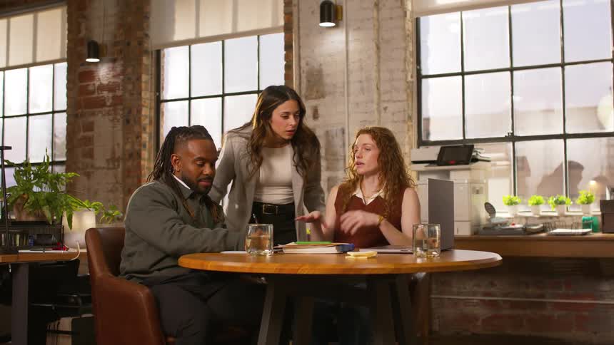 Three colleagues discuss documents and laptops at a round table inside an office with large windows and exposed brick walls