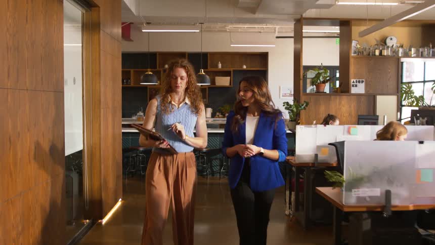 An office area is filled with workers in their cubicles at midday while two women walk down the hallway talking to each other