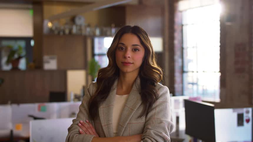 A woman with long wavy brown hair stands in a sunlit office with her arms crossed wearing a grey plaid blazershe begins by looking off to the side with a slight smile before turning her gaze directly toward the camera with a confident expression