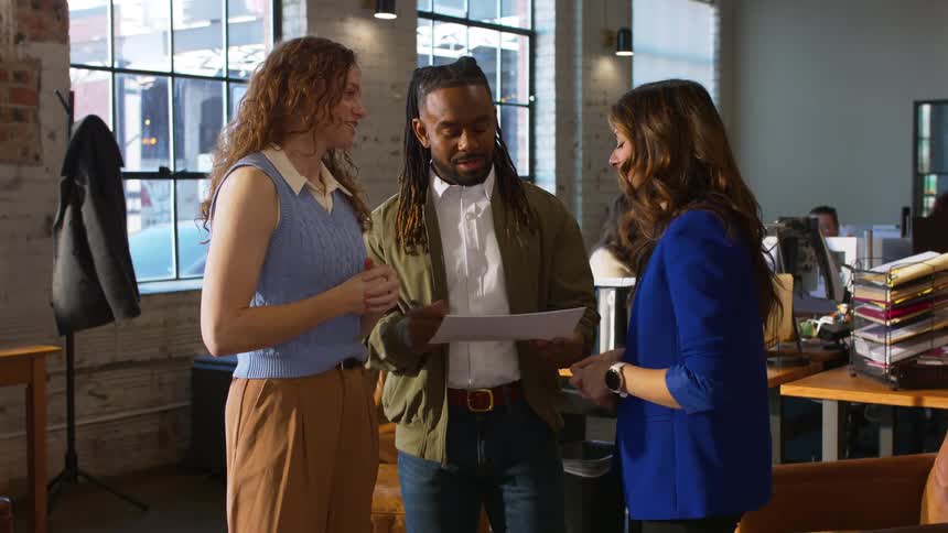 Two women and one man stand together in a bright office looking at a document while other colleagues work at desks in the background near large windows