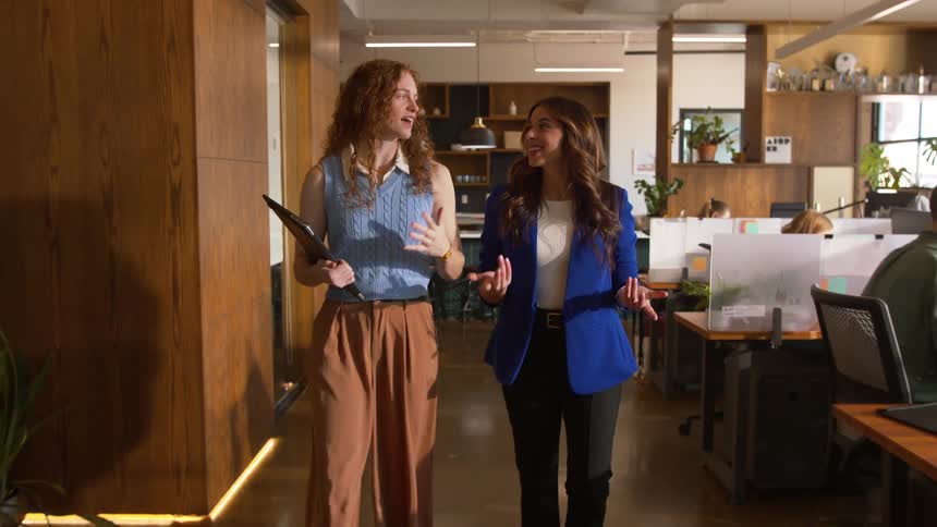 Two professional women walk through a modern openconcept office space while engaging in a friendly conversation the woman on the left carries a tablet and wears a blue sweater vest with tan trousers while the woman on the right wears a bright blue bla