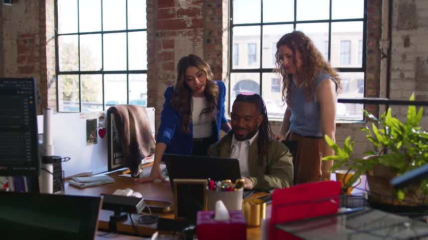 Three coworkers gathered at office desk with laptop plants and large windows in the background