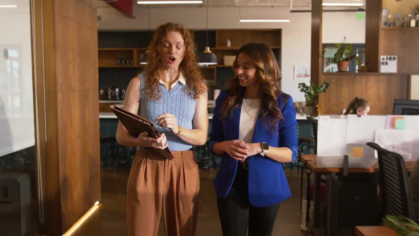 Two businesswomen walk through a modern office talking and smiling
