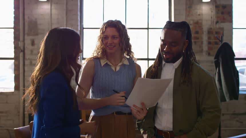 Three colleagues stand together in a sunlit office reviewing and discussing a document