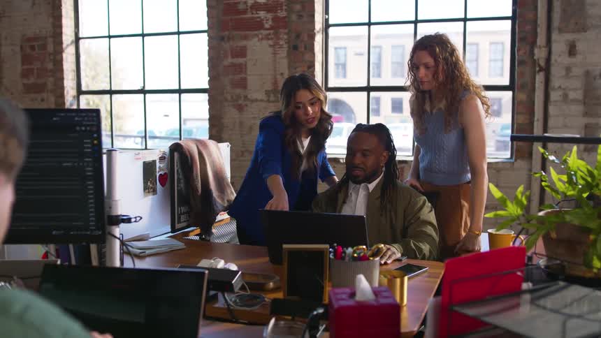 A woman walks over to join to coworkers at a cubicle to discuss work