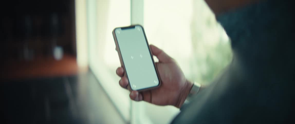 A man holds a smartphone while walking inside a bright room with a grey shirt and a watch on his wrist