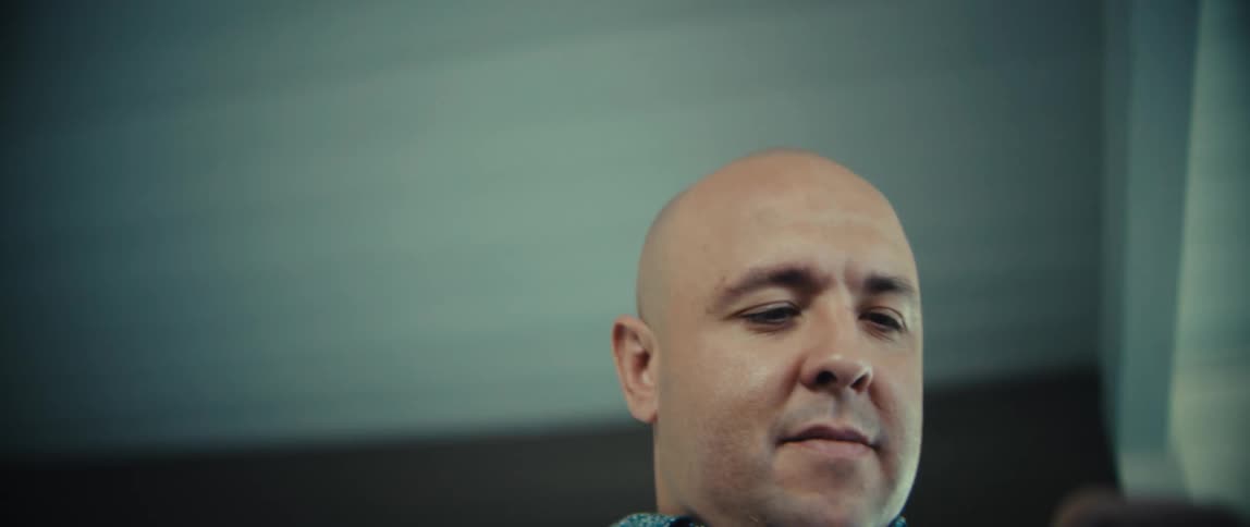A bald man in a blue patterned shirt looks down with a neutral expression while using his smartphone the camera captures a lowangle perspective as he navigates the device his face softly lit against a dark blurred background