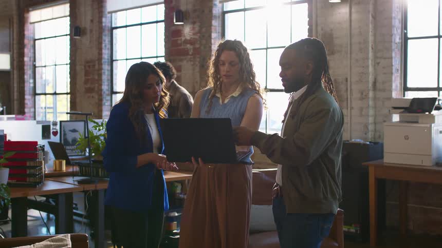 Three people review a laptop in an office with large windows in the background