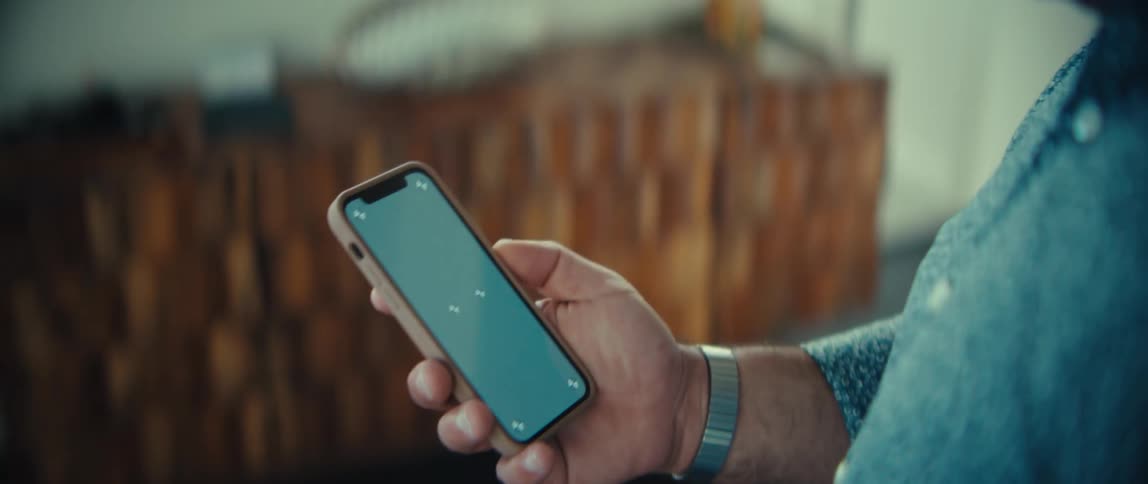 A man holds a smartphone featuring a green screen with tracking markers in a closeup shot while standing in a room with a wooden cabinet and blurred background elements