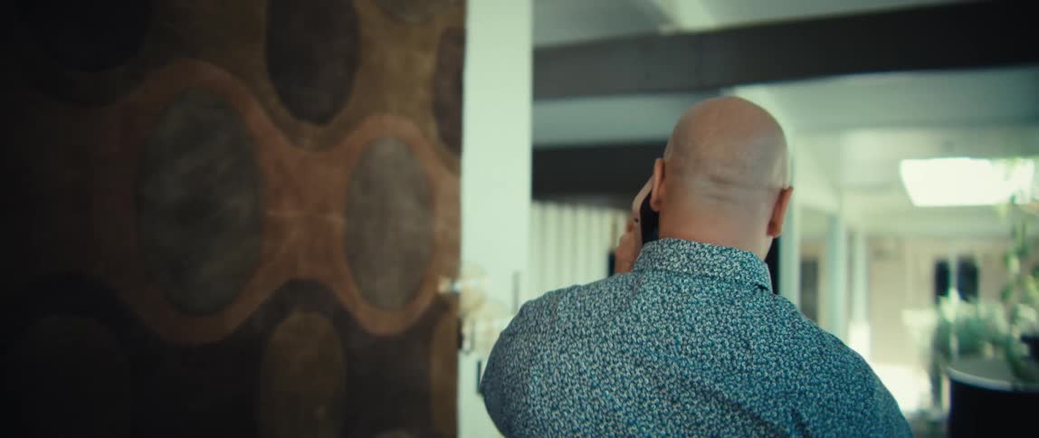 Man seen from behind talking on phone in spacious room with piano and large windows