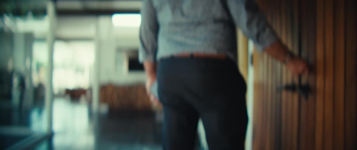 A man wearing a patterned shirt and dark trousers opens a wooden door and walks into a bright modern house during the day