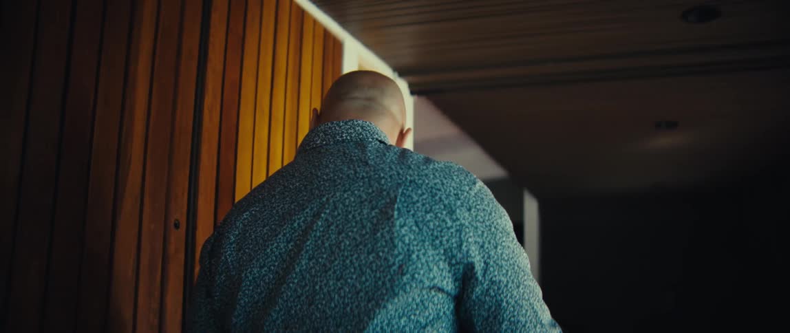 A man enters a home through a slatted wooden door while looking down at his smartphone he then walks further into the room and hangs a dark jacket on a wall hook