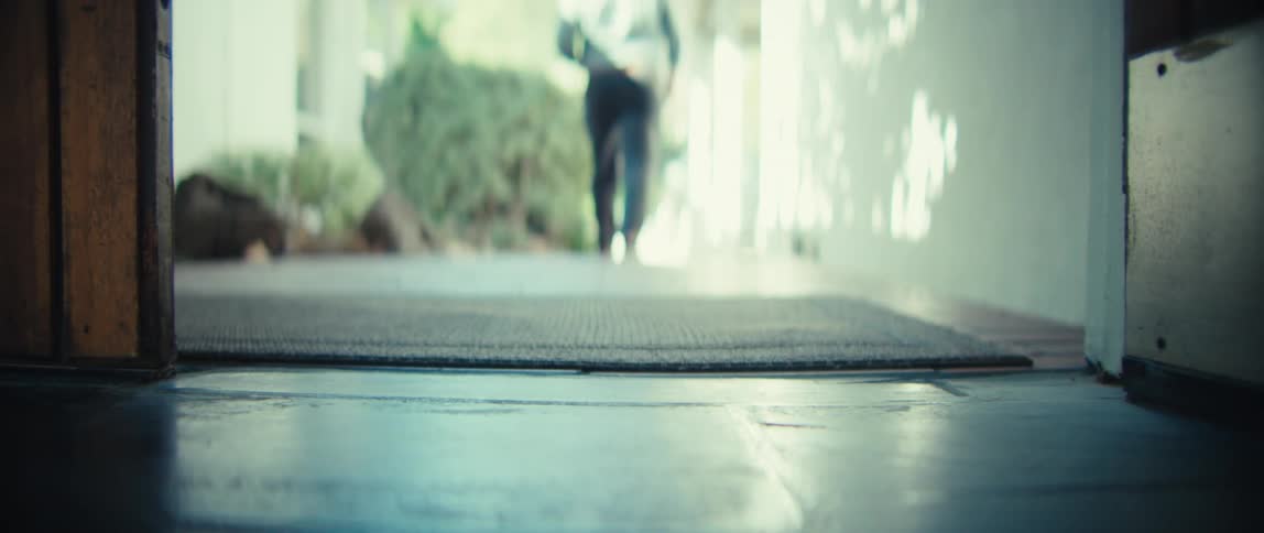 A person wearing black pants and dark shoes walks through an open wooden door and across a floor mat moving away from the camera into a bright outdoor patio area with green plants in the background