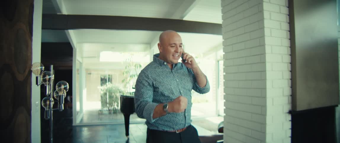 A bald man wearing a patterned shirt walks through a bright modern house while talking on a mobile phone and smiling during the day