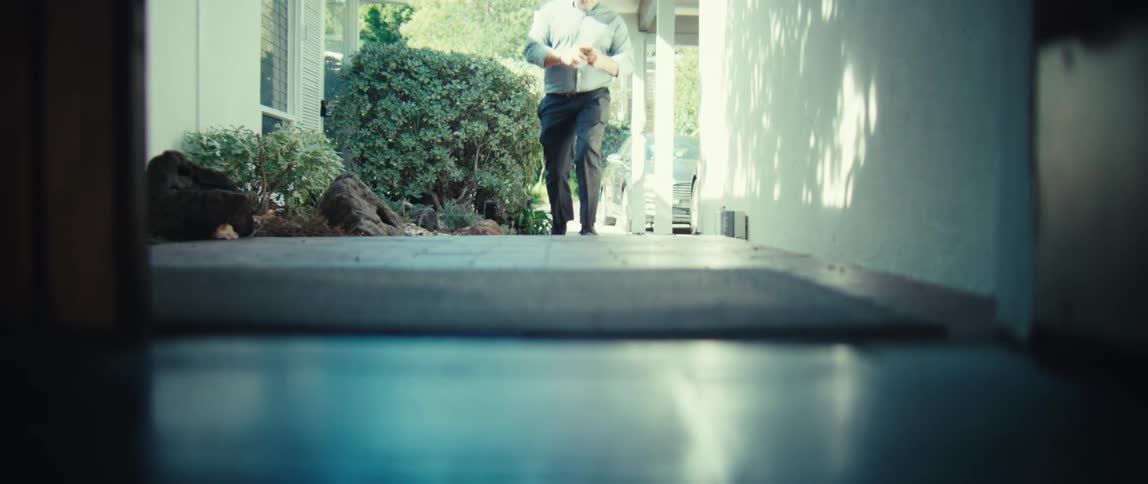 A bald man wearing a light blue shirt and dark pants walks down a bright outdoor walkway toward a residential entrance while looking at his phone