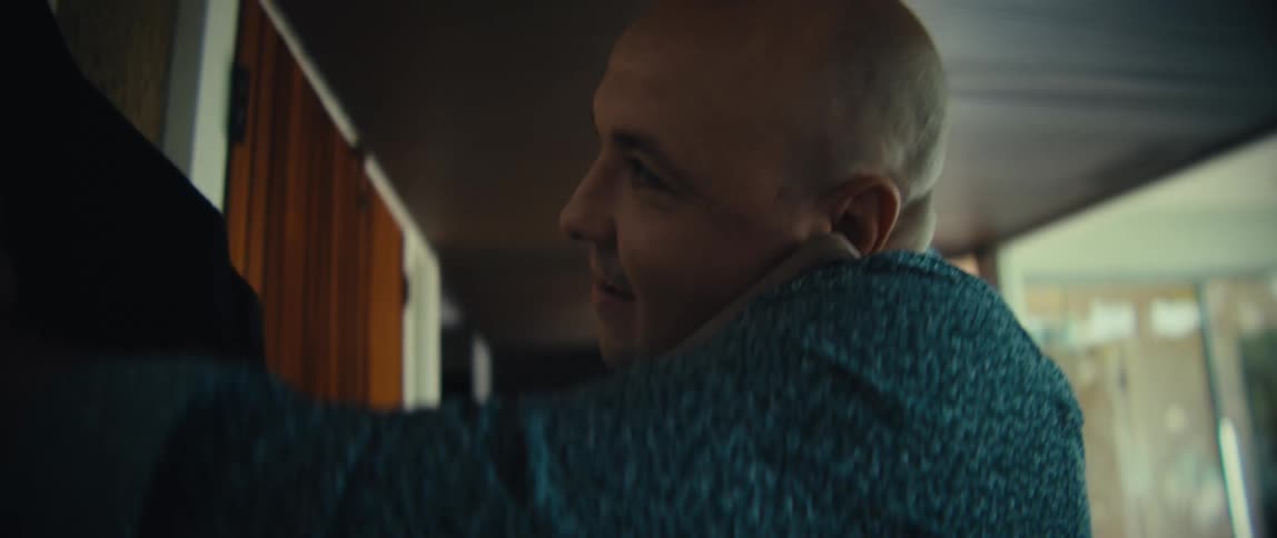 A bald man wearing a blue patterned shirt talks on a mobile phone while entering a bright modern house and hangs his black jacket on a wall hook before walking further inside with a happy expression