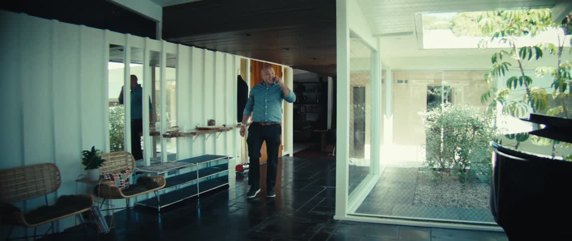 A bald man in a blue patterned shirt walks through a modern hallway while talking on a smartphone