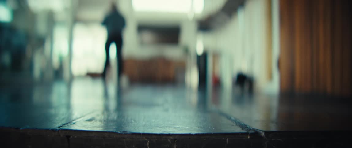 A close view of dark shoes and black trousers as a man walks across a dark tiled floor inside a bright house with wooden walls and large windows in the background