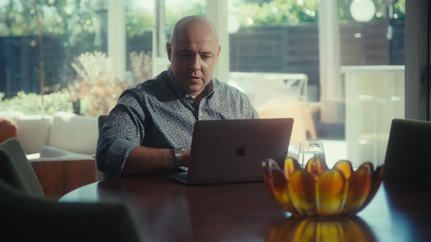 A bald man in a grey shirt sits at a wooden dining table and types on a laptop near a decorative glass bowl while bright natural light filters through large windows in the background of a modern home interior