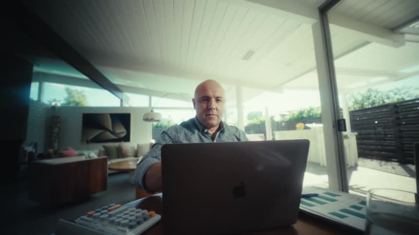 The video shows a man in a patterned blue shirt setting up his workspace in a modern sunlit room after adjusting his laptop screen he settles in to begin working at a wooden desk equipped with a calculator and various documents