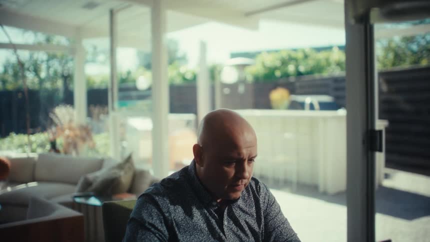 A bald man wearing a patterned blue shirt sits at a table and works on a laptop in a bright modern house with large windows and hanging lights