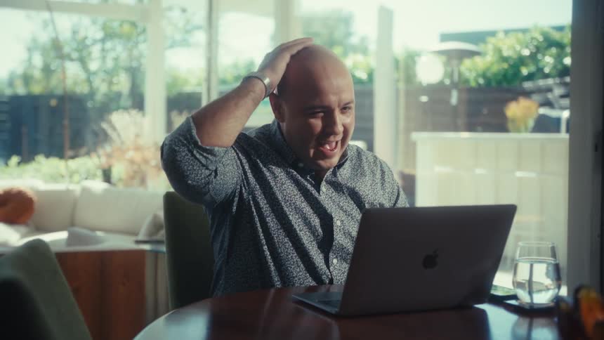 Man seated at a table holds his head while looking at an open laptop